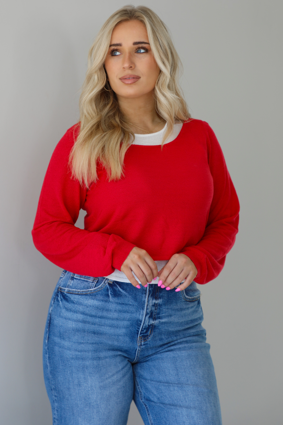 Woman wearing a red and white layered appearance top with dark wash denim jeans. The photo is taken indoors against a white wall.