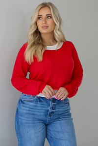 Woman wearing a red and white layered appearance top with dark wash denim jeans. The photo is taken indoors against a white wall.