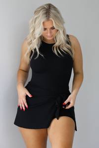 Woman wearing a black athleisure romper that has a wrap-style skirt detail that wraps around and ties in the front on the left side. The photo is taken indoors against a white wall with white marbled flooring. 