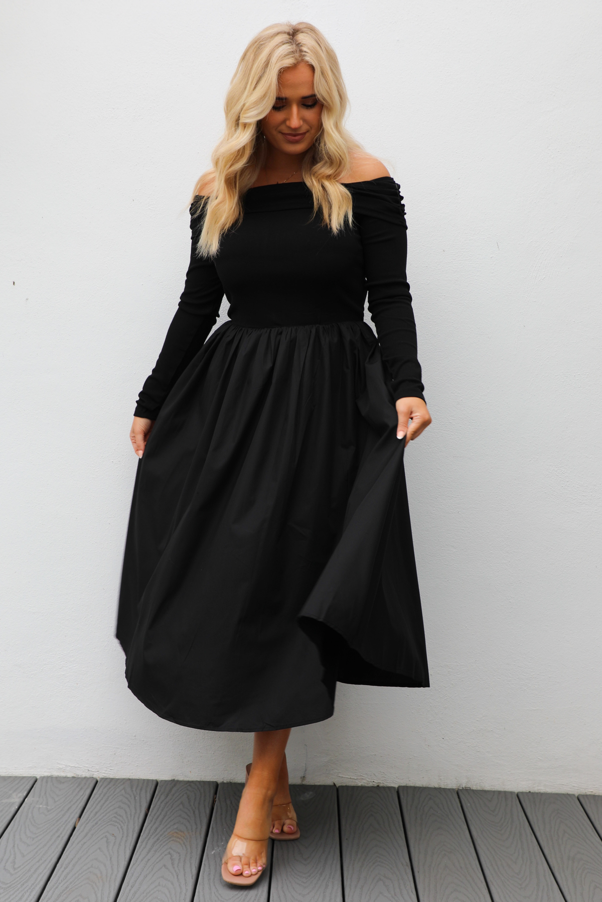 Woman wearing a black off the shoulder, long sleeved midi length dress with a pair of clear strapped nude heels. The photo is taken outdoors against a white wall.