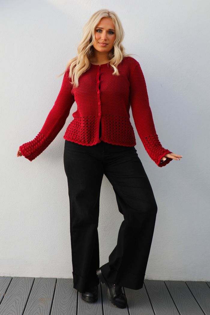 Woman model wearing a burgundy knit cardigan with button closures in the front and black denim jeans. The photo is taken outdoors against a white wall. 