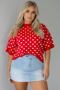 Woman wearing a red knit top with a white polka dot print pattern throughout with a high waisted, light-wash denim skort. The photo is taken indoors against a white wall.