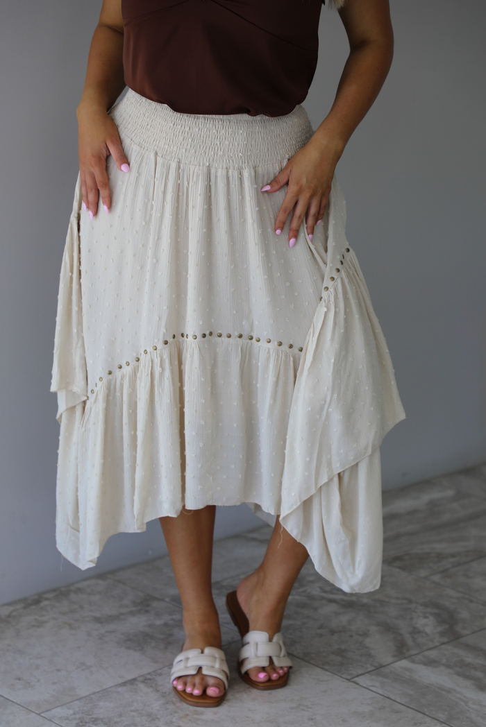 Woman wearing a sand colored midi skirt featuring a smocked waistline, a polka dot textured material, a studded detail, and a midi length. The photo is taken indoors against a white wall with white marble flooring. 