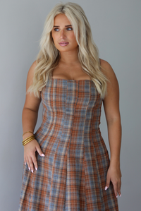 A woman wearing a brown and blue plaid midi dress with thick straps, a square neckline, fitted bodice that starts flaring out at the hips and ends about mid-shin, with a flowy skirt, standing indoors against a white wall. The photo shows a closeup of the bodice of the dress. 