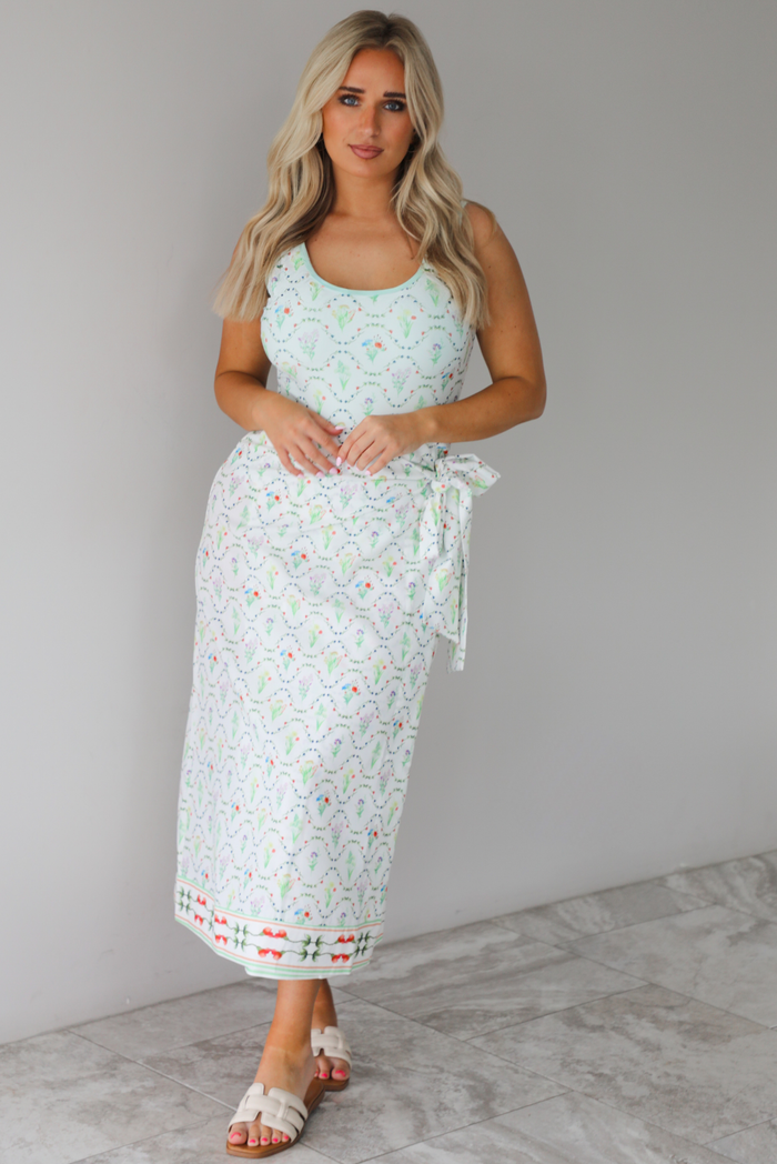 Woman wearing an off white swimsuit & coverup skirt set that has a floral print pattern throughout. The photo is taken indoors against a white wall with white marble flooring. 