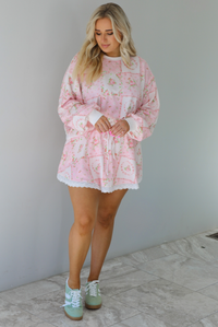 Woman wearing a two piece set featuring a pink floral set that has an oversized long sleeved top and high waisted skirt. The photo is taken indoors against a white wall with white marble flooring. 