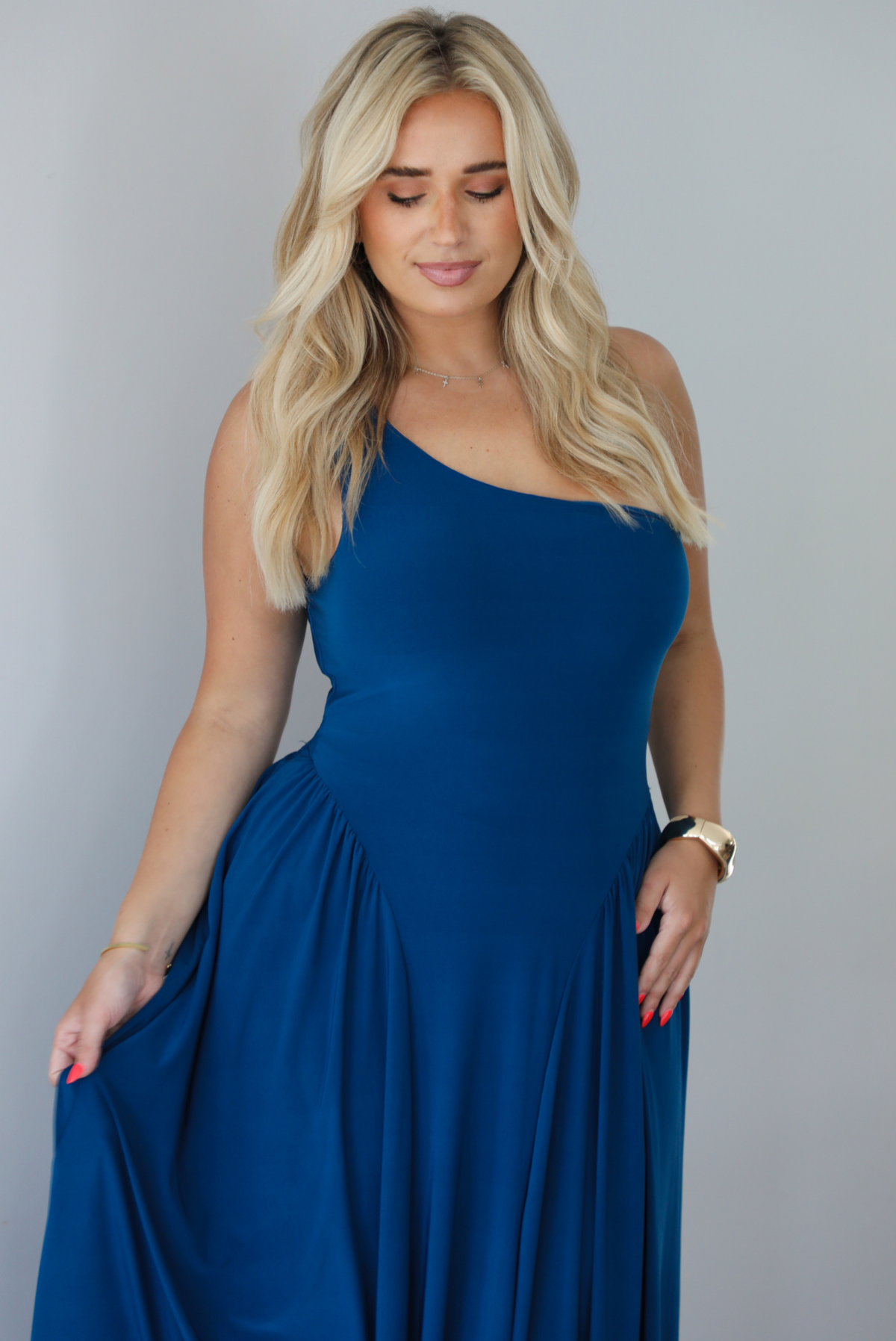 Woman wearing a blue one strap maxi dress indoors against a white wall. The photo shows a closeup of the bodice of the dress. 