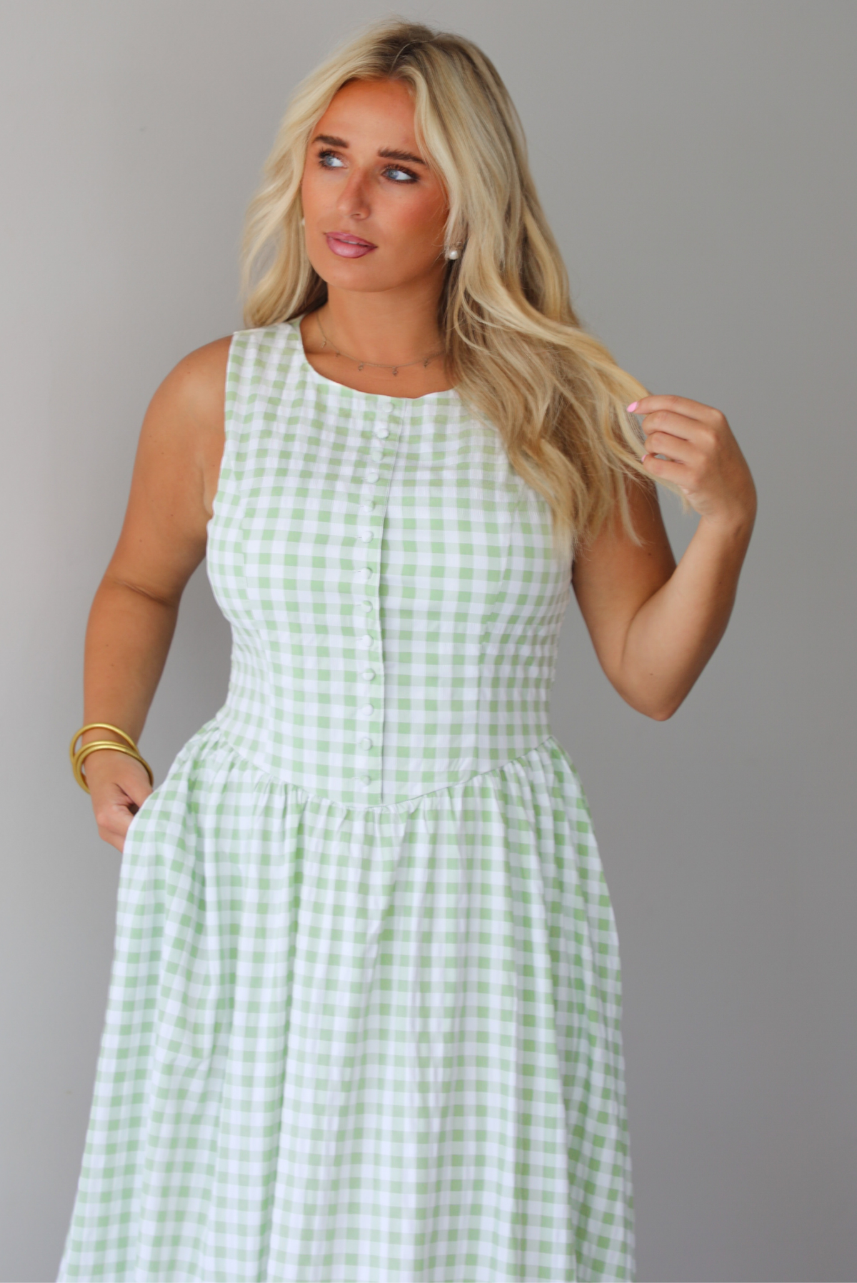 A woman wearing a light green and white gingham maxi dress with a drop waist bodice and side pockets, standing indoors against a white wall. It shows a closeup o the bodice of the dress, which has non-functional button closures in the front. 