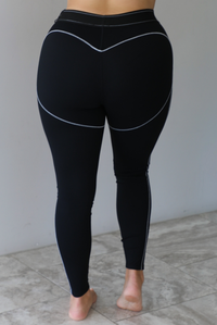 A woman modeling a matching black activewear set consisting of a sports bra and high-waisted leggings with thin white contrast stitching, standing indoors against a white wall with white marble flooring. The photo shows a closeup of the back of the leggings. 