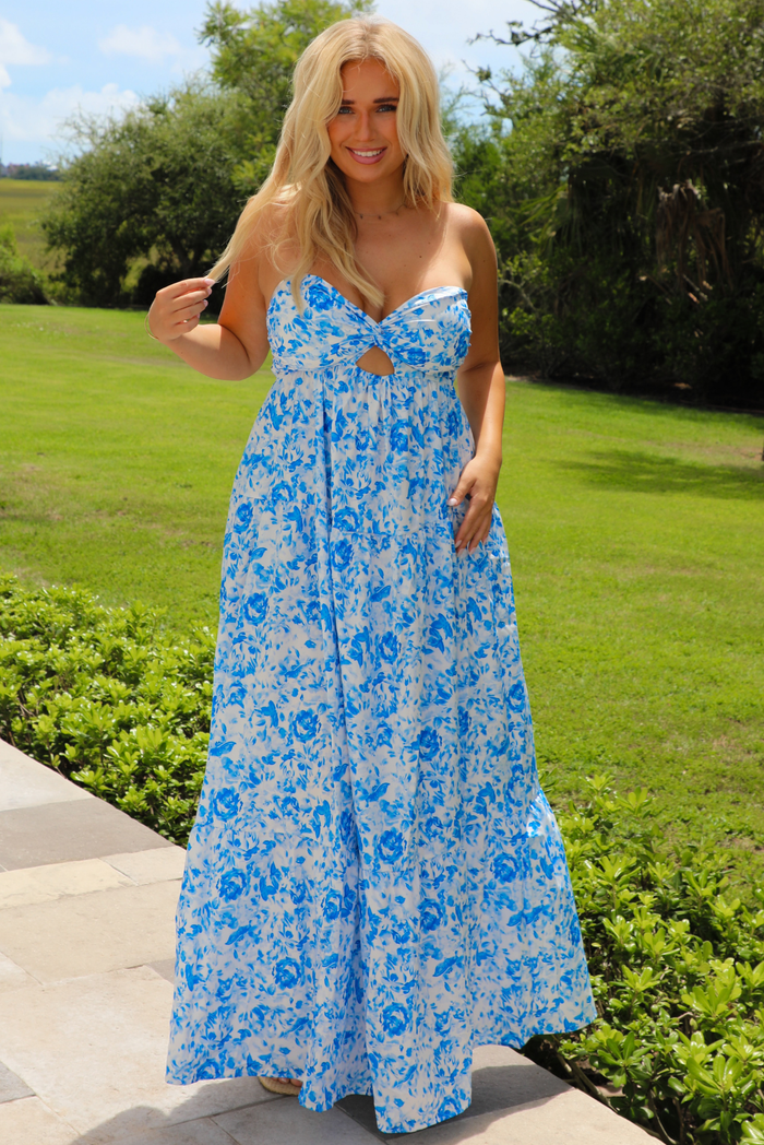 A woman wearing a strapless blue and white floral maxi dress with a keyhole cutout at the bust, standing outdoors on a stone path with a green lawn and trees in the background.