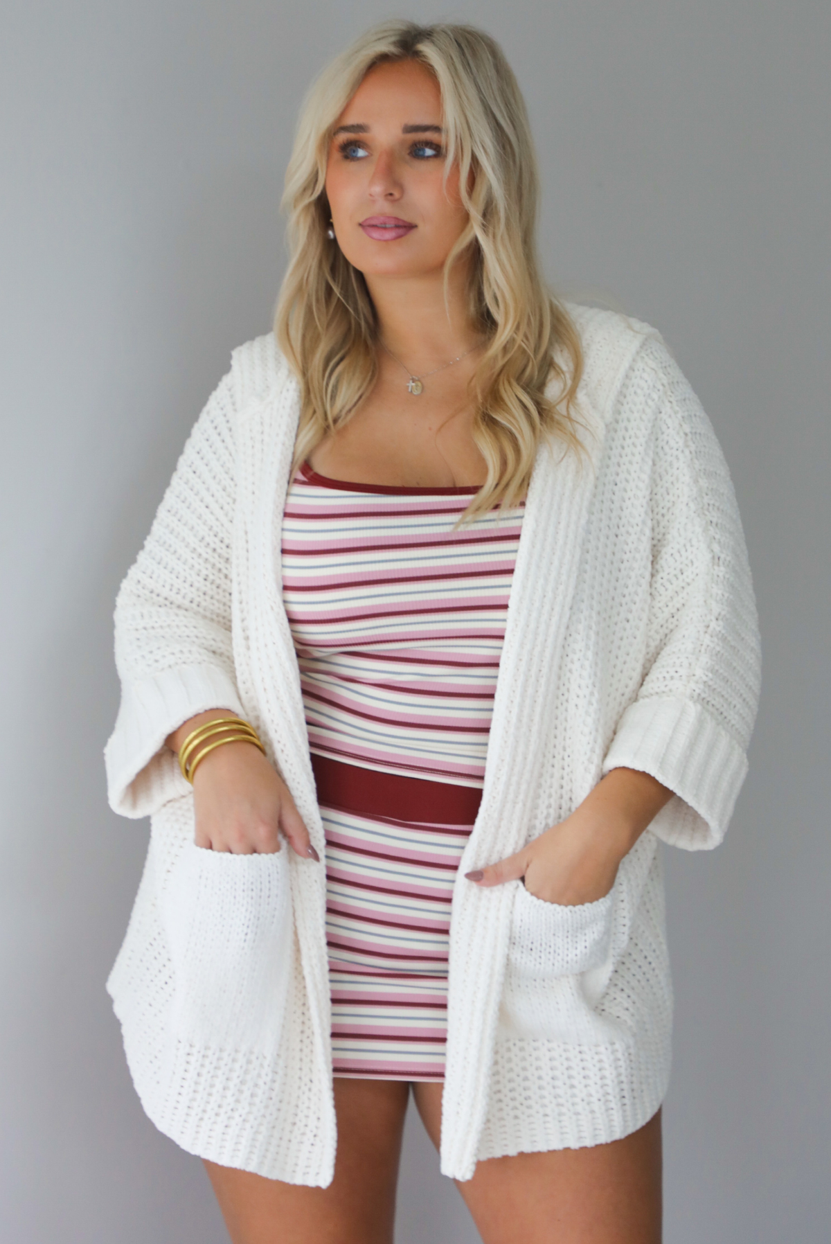 A woman wearing a cream colored oversized cardigan worn over a striped two piece set, standing indoors against a white wall.