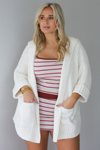 A woman wearing a cream colored oversized cardigan worn over a striped two piece set, standing indoors against a white wall.
