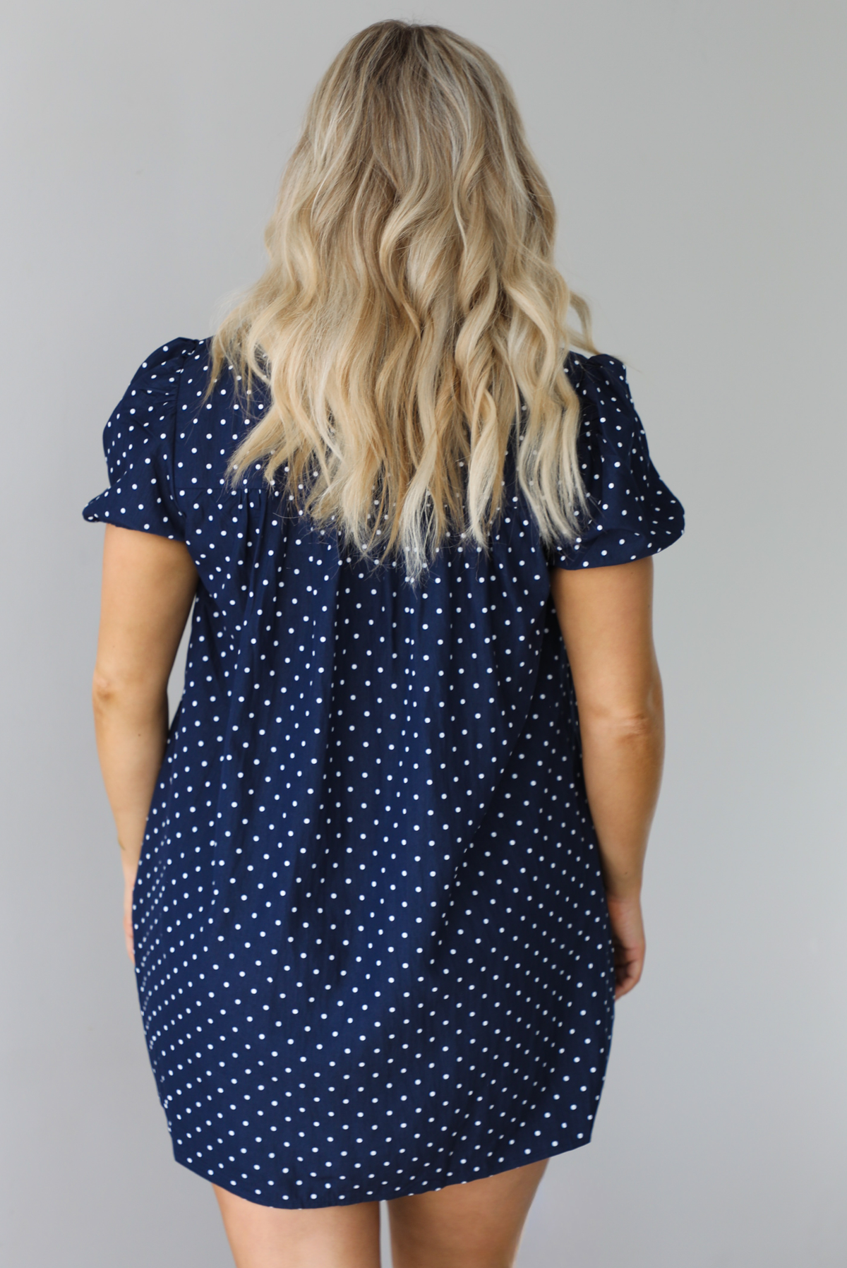 Woman wearing a navy baby-doll cut dress with a white polkadot print pattern throughout. The dress has puffed short sleeves and functional pockets on either side. The photo shows the back of the dress. The photo is taken indoors against a white wall.