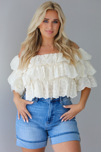 Woman wearing a cream off the shoulder, ruffled eyelet cropped top with medium wash denim shorts indoors against a white wall.
