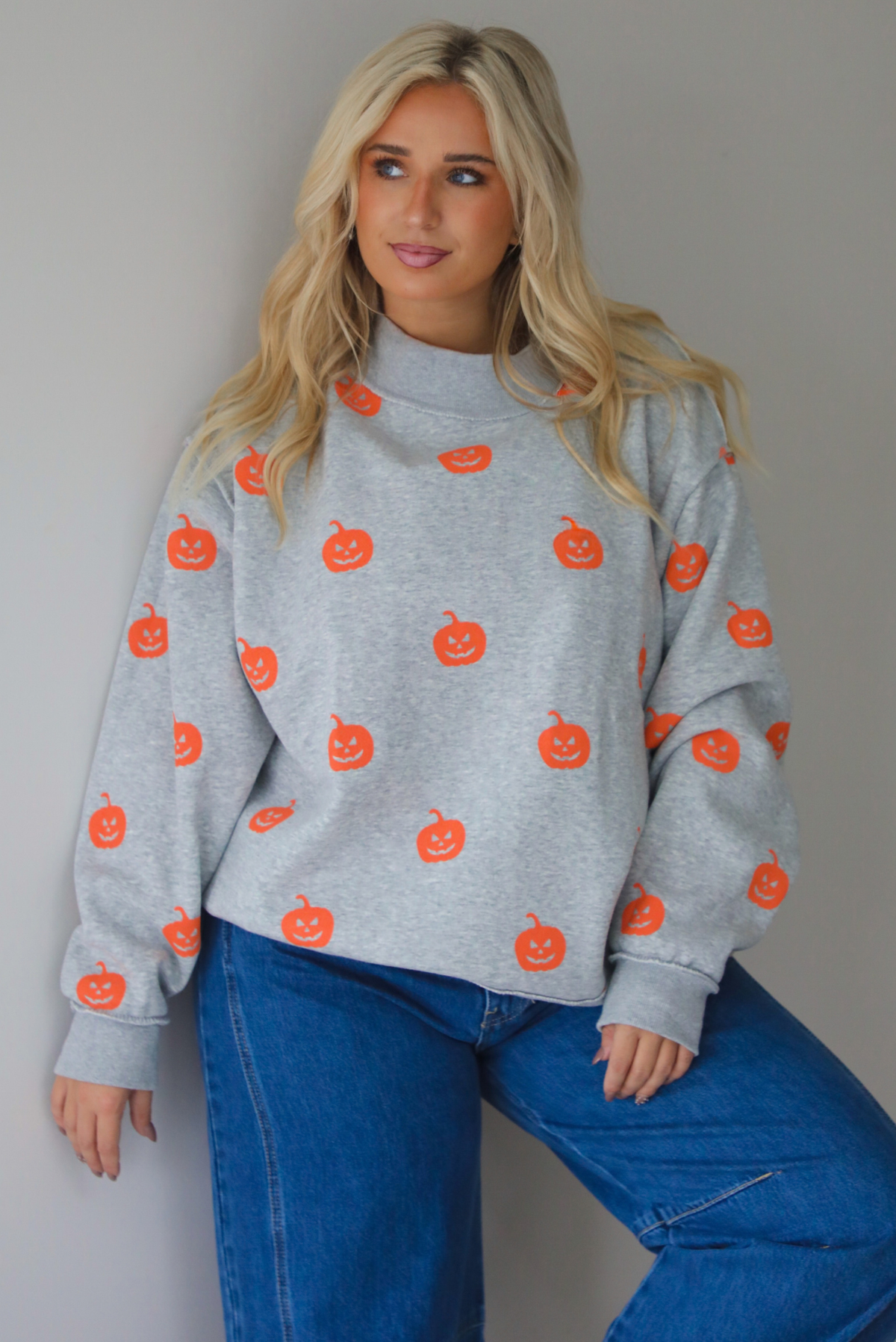 A woman wearing a heather grey oversized sweatshirt that has a repeating jack-o-lantern design throughout, paired with dark wash denim jeans, standing indoors against a white wall.