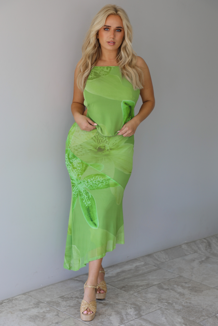 Woman wearing a green tropical print set featuring a backless tank and high waisted, asymmetrical hem midi skirt indoors against a white wall with white marble flooring. 