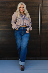 Woman model wearing a mocha brown cardigan that has grey stitching details throughout. The cardigan has a cropped cut, is oversized, and has functional button closures in the front. She's wearing the cardigan with dark denim jeans and brown shoes. The photo is taken against a dark brown wooden door outdoors. 