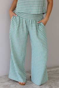 Woman wearing a mint and white gingham set with an oversized top and flared hem pants indoors against a white wall with white marble flooring. The photo shows a closeup of the pants, which have a smocked waistline and functional pockets on either side. 