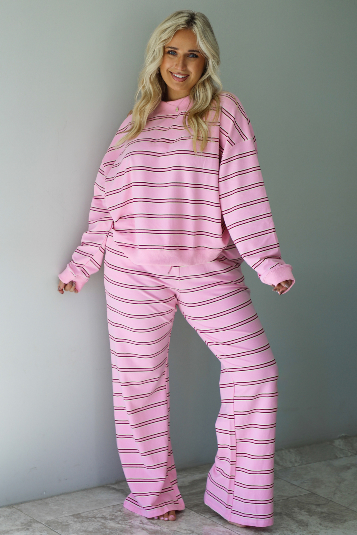 A woman wearing a pink two piece sweat set featuring a maroon and white striped design on a sweatshirt and high waisted, wide leg pants, standing indoors against a white wall with white marble flooring. 