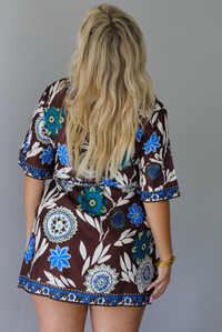 A woman shown from behind modeling a brown short-sleeve wrap mini dress with a colorful blue, teal, and white floral pattern, paired with beige knee-high boots, standing indoors against a white wall.
