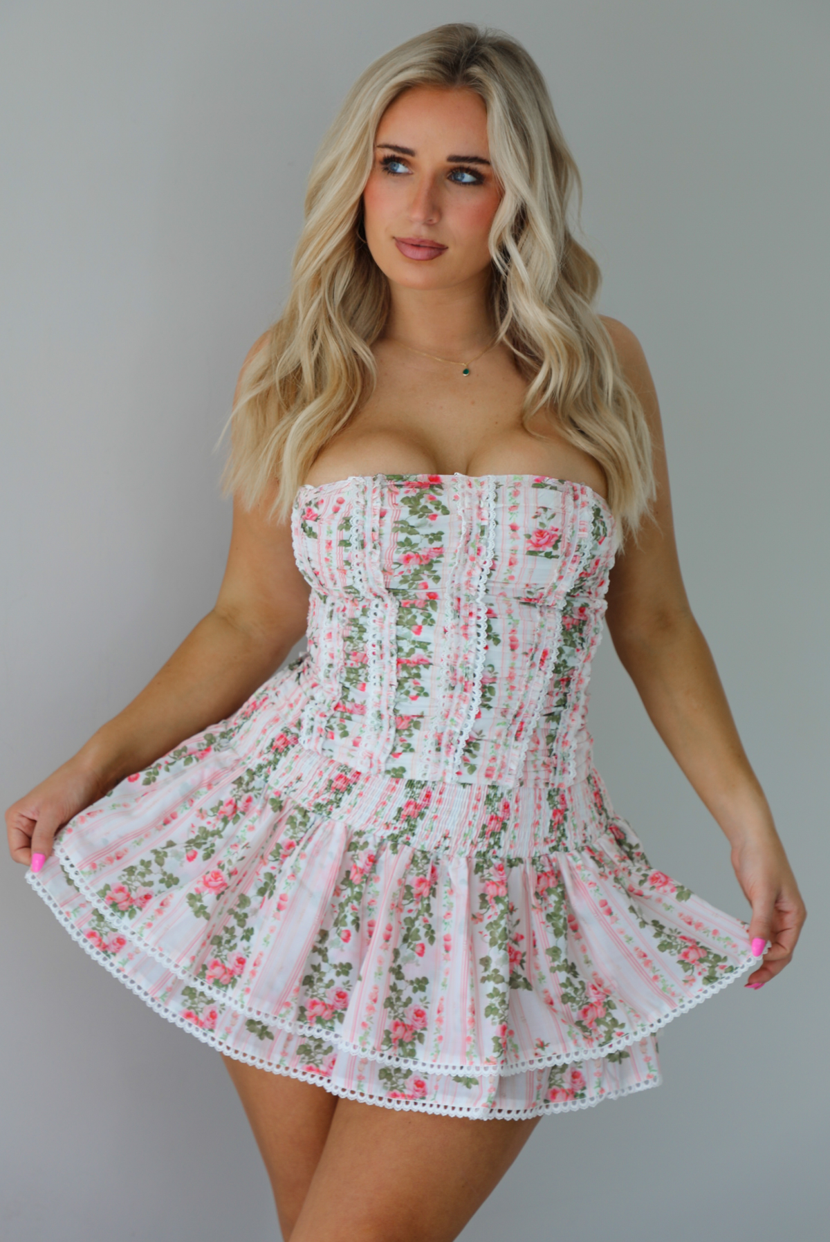 Woman wearing a white and pink floral two piece set featuring a strapless corset style top and an a ruffled skirt. The photo is taken indoors against a white wall.