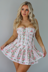 Woman wearing a white and pink floral two piece set featuring a strapless corset style top and an a ruffled skirt. The photo is taken indoors against a white wall.
