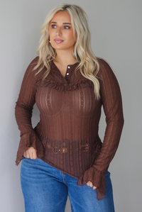 A woman wearing a sheer brown lace long sleeved top with a quarter-button closure detail and a ruffled detail at the top of the bodice, untucked to show the full length,  paired with dark wash denim with a black belt, standing indoors against a white wall.