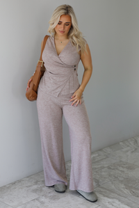 Woman wearing a taupe colored two piece set featuring a vest-appearance top with non-functional button closures on the left side and high waisted pants. It's paired with a tan colored vegan leather shoulder bag. The photo is taken indoors against a white wall with white marble flooring. 