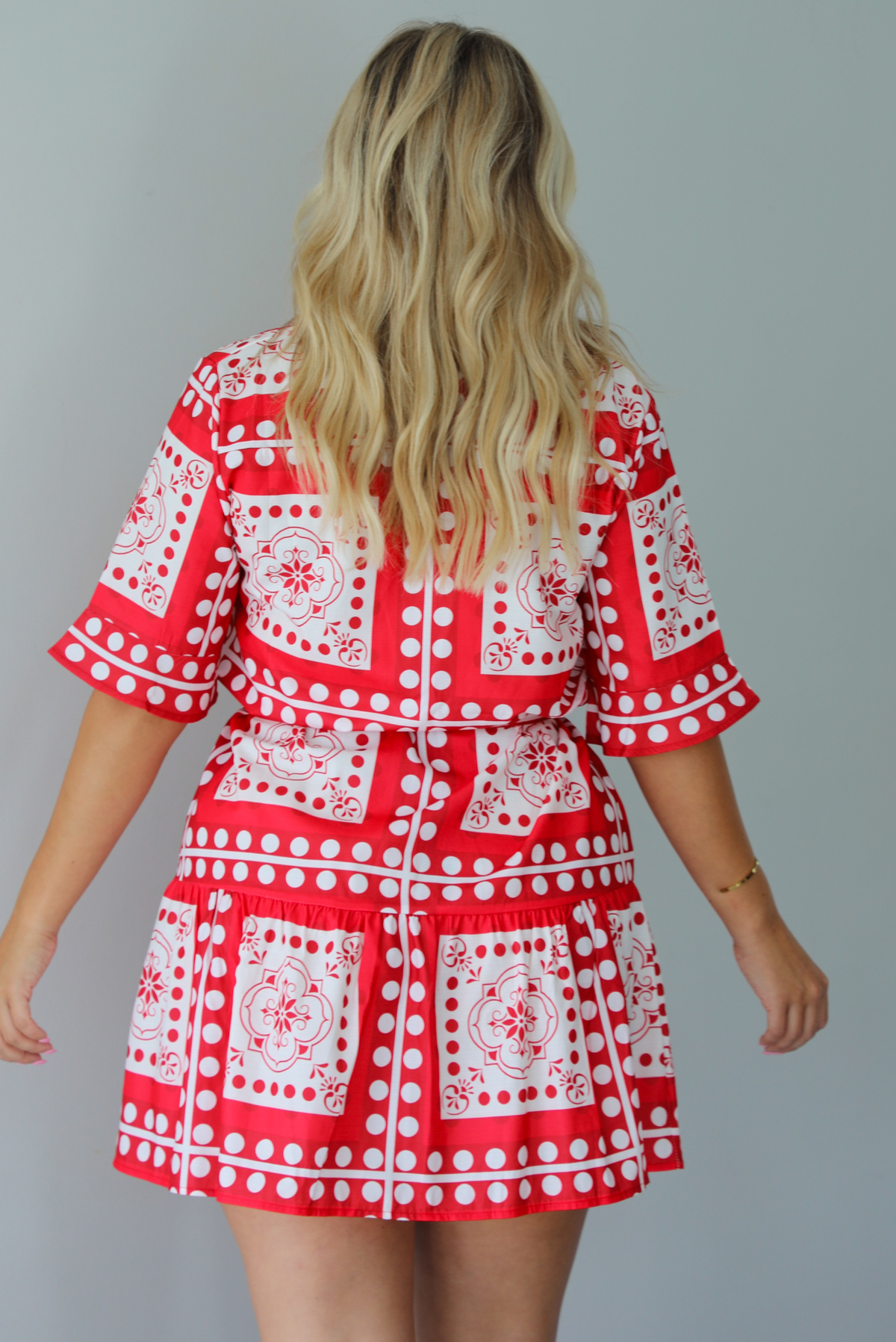 A woman shown from behind modeling a red and white patterned short-sleeve mini dress with button closures along the front and a tie waist, standing indoors against a white wall.