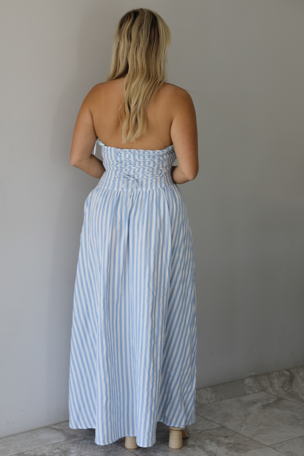 Woman wearing a blue and white striped strapless maxi dress indoors against a white wall with white marble flooring. The photo shows the back of the dress, which has a corset-like lace up design.
