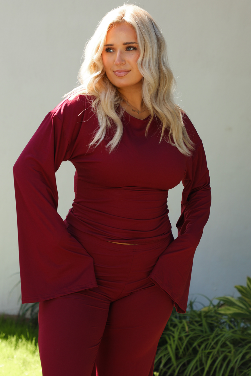 Close up of a woman model wearing a burgundy wine colored two piece set outdoors. The photo shows the front of the top of the set. The top features a slightly cropped, long sleeved design that has flared sleeves, a fitted design, and a boat neckline. 