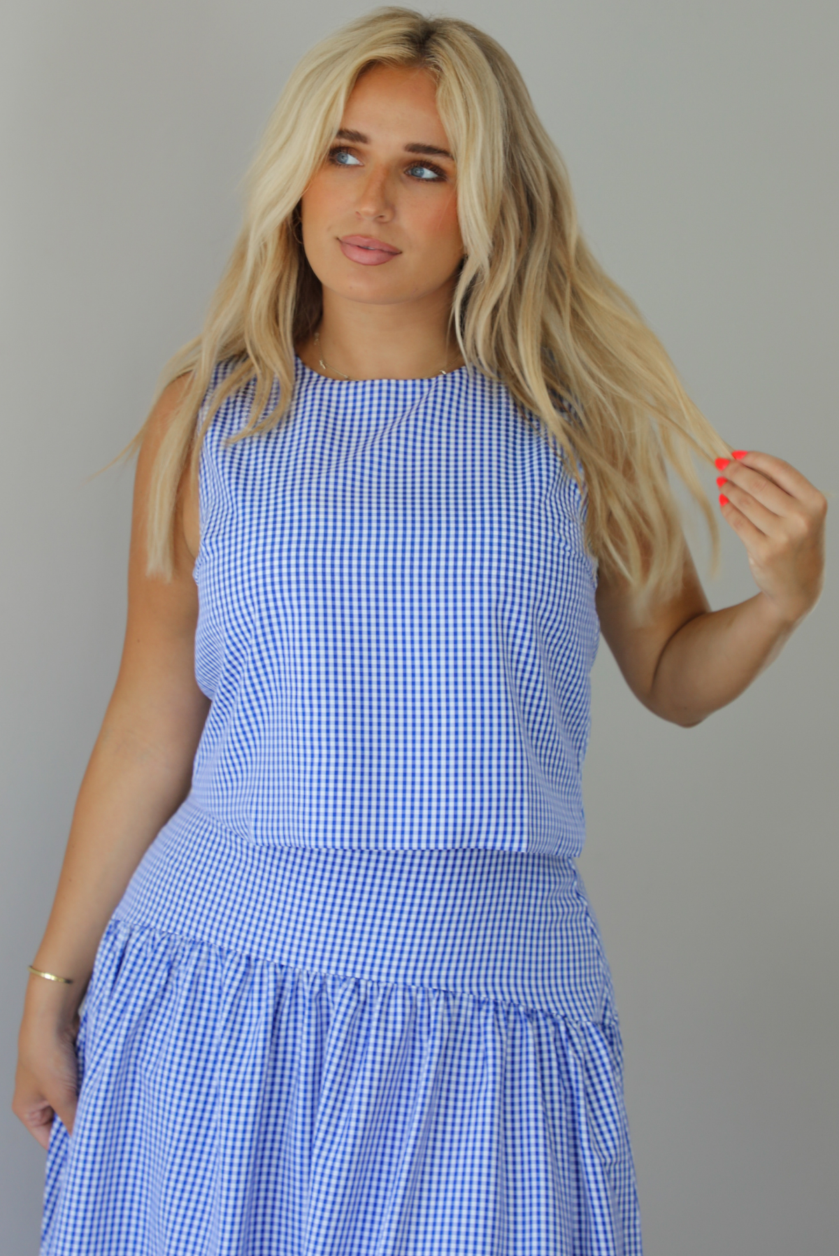 Woman wearing a blue and white gingham two piece set featuring a sleeveless top and high waisted, maxi length skirt indoors against a white wall The photo shows a closeup of the top. 