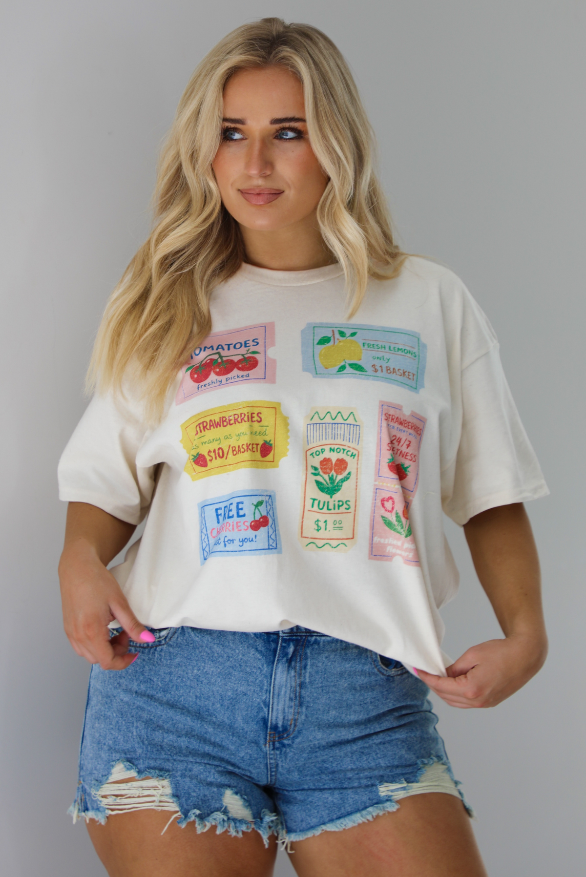 Woman wearing a white relaxed fit graphic tee that has various tickets on it with different fruits and flowers on them. The tee is tucked into a pair of distressed denim shorts. The photo is taken indoors against a white wall.