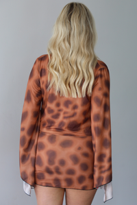 Woman wearing a brown and ruse mini dress that has a very low slouched cowl-neck design, long sleeves, and is fitted at the hips indoors against a white wall. The photo shows the back of the dress. 