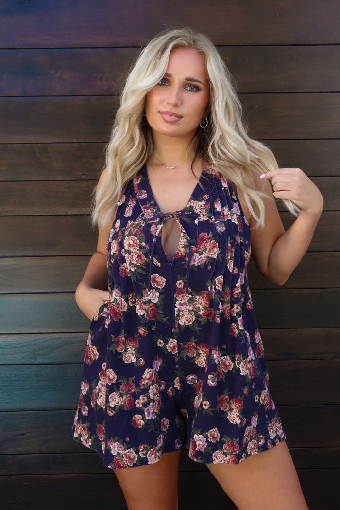 Woman model wearing a navy with pink and muted red floral print patterned denim romper that has a v-neckline with a tie closure in the center, related details along the bodice, functional pockets on either side, and a relaxed fit. The photo is taken outdoors against a dark brown wooden door.