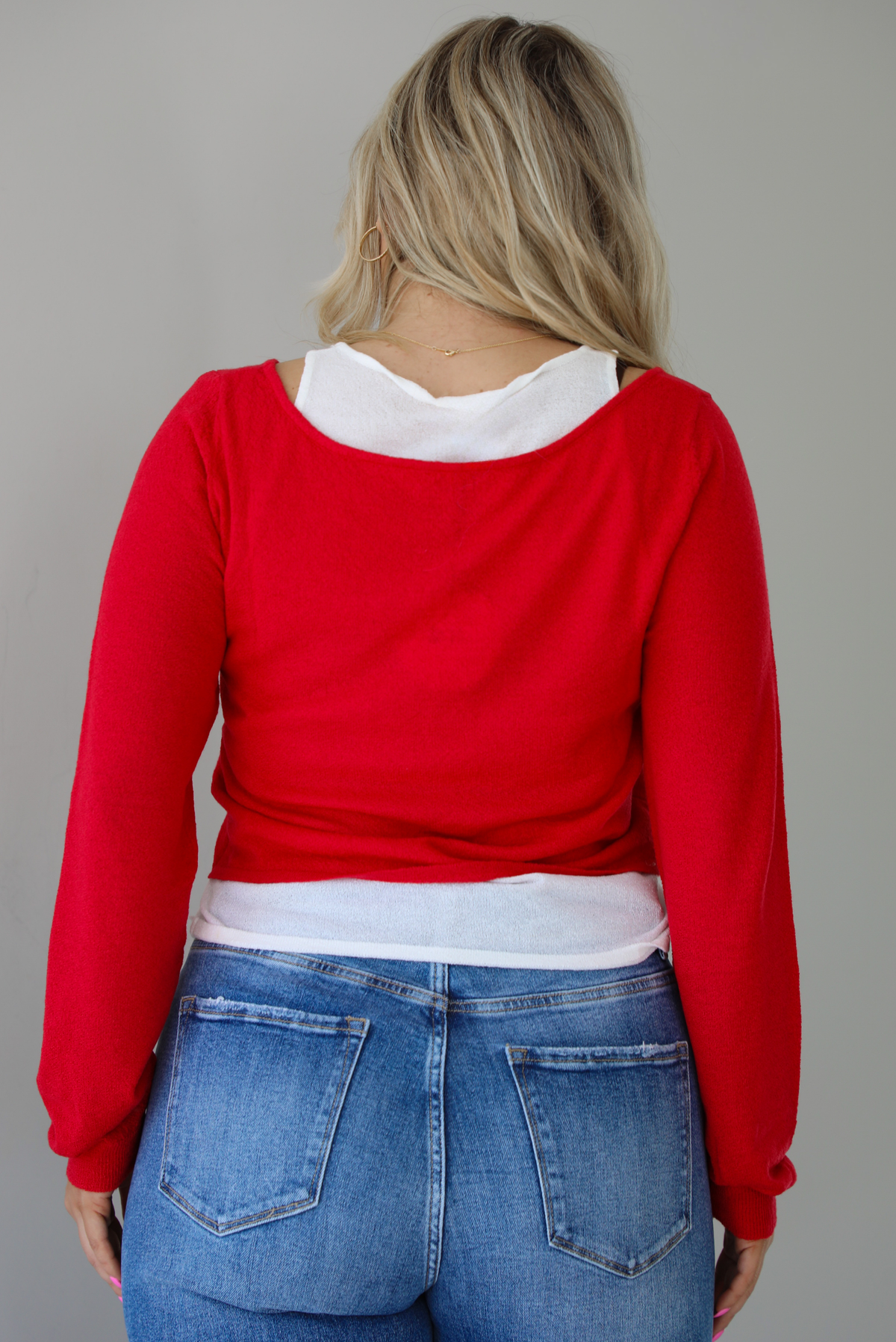 Woman wearing a red and white layered appearance top with dark wash denim jeans. The photo is taken indoors against a white wall. The photo shows the back of the top.
