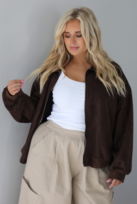 A woman wearing a brown suede jacket with a white tank and sand colored baggy pants, standing indoors against a white wall. The photo shows a closeup of the jacket, which has a zipper closure in the front and cinched details at the wrists. 