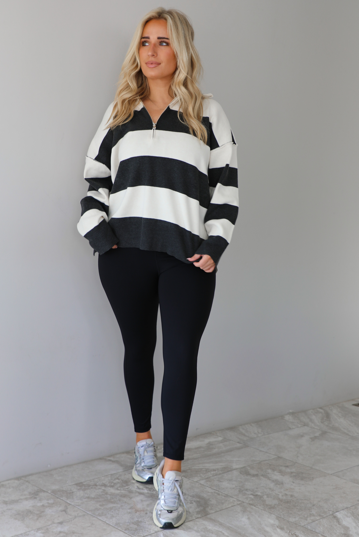 Woman wearing a charcoal and white striped quarter-zip sweater with a collared neckline, raw seam details, and a relaxed fit. It's paired with a fitted black jumpsuit and silver sneakers. The photo is taken indoors against a white wall with white marble flooring.