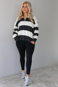 Woman wearing a charcoal and white striped quarter-zip sweater with a collared neckline, raw seam details, and a relaxed fit. It's paired with a fitted black jumpsuit and silver sneakers. The photo is taken indoors against a white wall with white marble flooring.