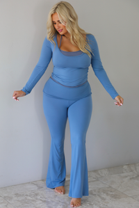 A woman modeling a matching two-piece lounge set in a dusty blue color, featuring a long-sleeve scoop-neck top with a connected darker blue halter neck detail and matching high-waisted flared pants, standing indoors against a white wall with white marble flooring. 