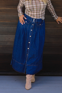 A woman wearing a long blue denim button-front skirt with a plaid long-sleeve top and beige ankle boots, standing against a dark wooden door. 