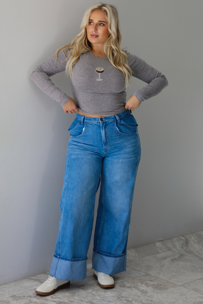 A woman wearing a striped, cropped long sleeved top with a beaded espresso martini on the front with medium wash denim jeans, standing indoors against a white wall with white marble flooring 