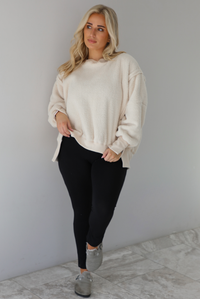 Woman wearing a sherpa sand oversized sweater with a fitted black jumpsuit underneath and grey slide on shoes. The photo is taken indoors against a white wall with white marble flooring. 