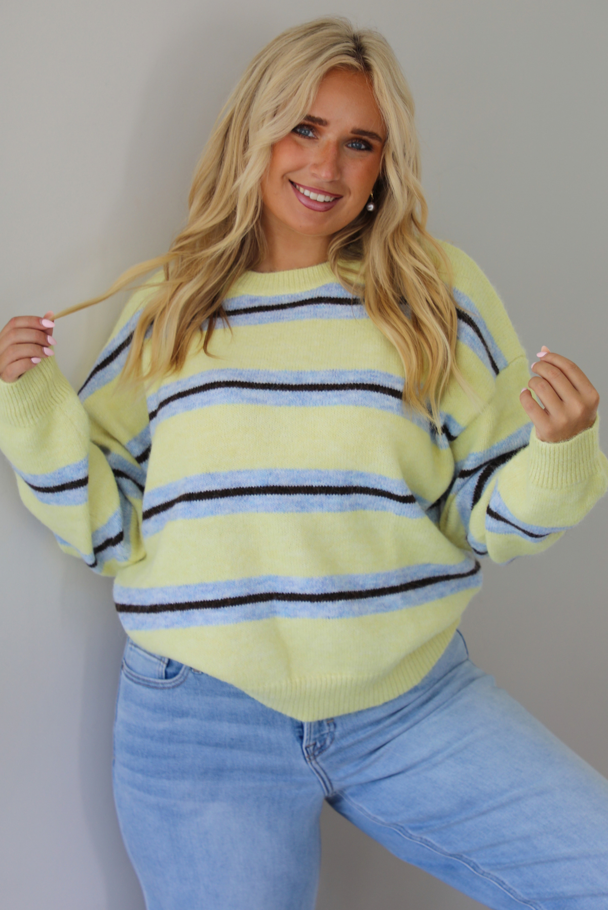 Woman wearing a pastel yellow, blue, and dark brown striped sweater, paired with light wash denim jeans that have a wide leg cut with a cuffed hem, standing indoors against a white wall.