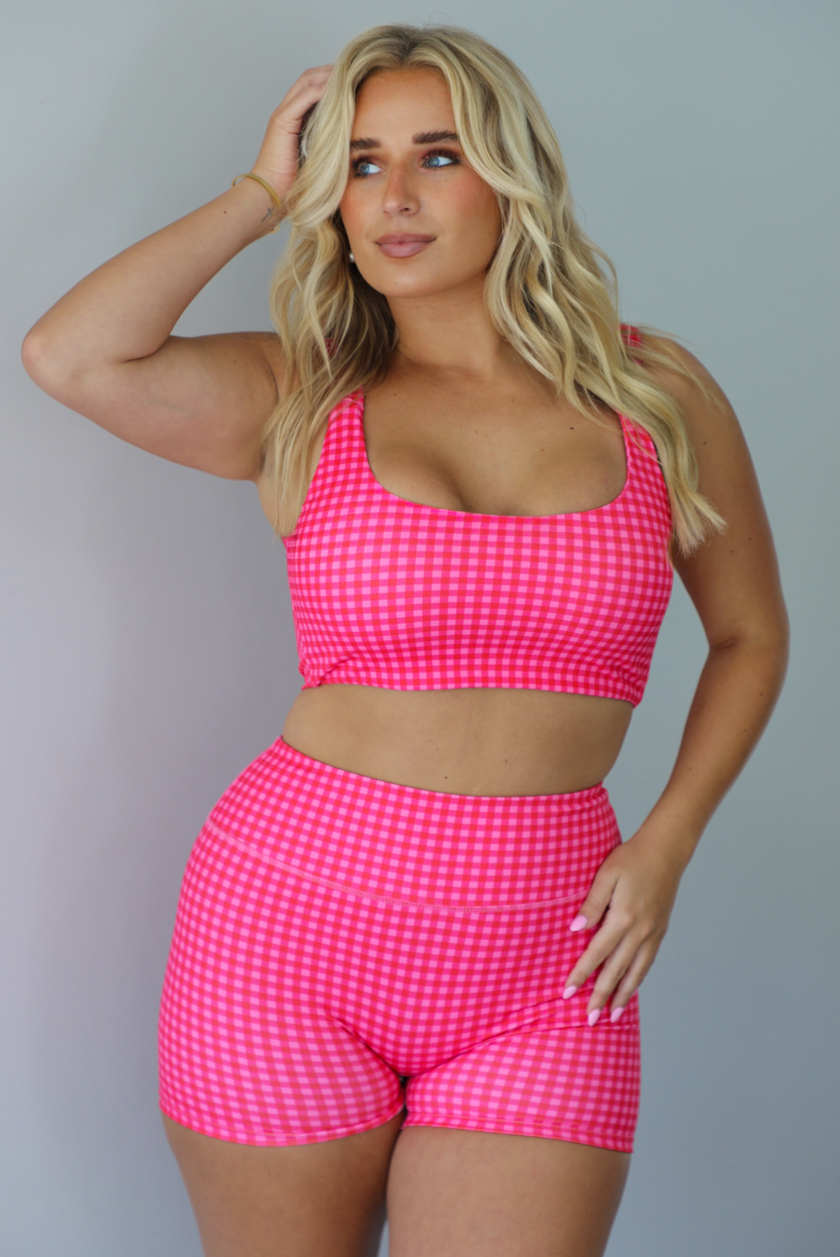 A woman wearing a red and hot pink gingham two-piece activewear set consisting of a crop top and matching shorts, standing indoors against a white wall.