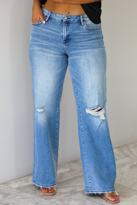 Woman wearing a pair of medium-wash, mid-rise denim jeans that have distressed details in the front and a flared, boot-cut-like hem. The photo is taken indoors against a white wall with white marble flooring. 