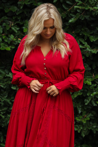 Woman wearing a red maxi length dress that has a v-neckline with button closures at the bodice, a cinched with elastic waistline, long sleeves, and a flowy skirt. The photo is taken outdoors against greenery.  The photo shows a closeup of the bodice, sleeves, and waistline. 