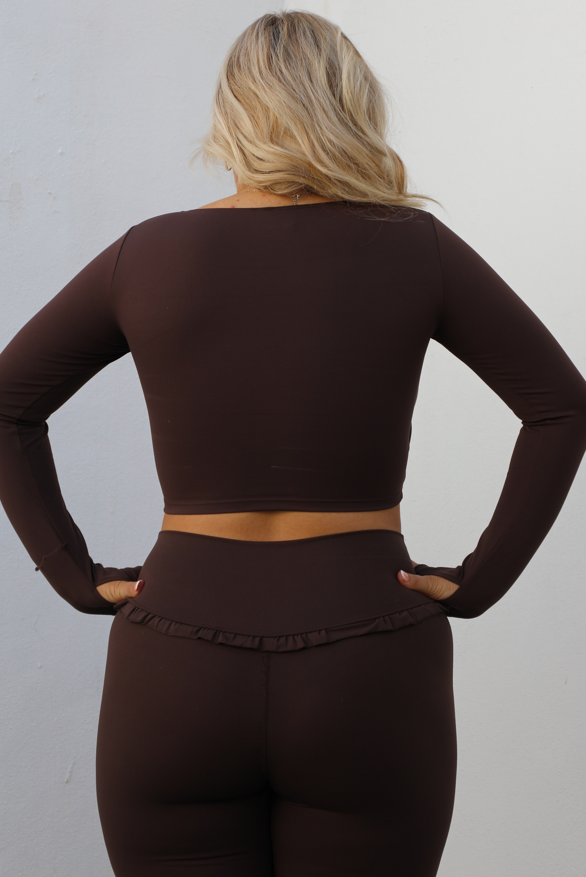 Woman model wearing a brown two-piece athleisure set. The top is cropped and the pants are high waisted. The photo is taken outdoors against a white wall. The photo showcases the back of the top and the upper half of the pants. 