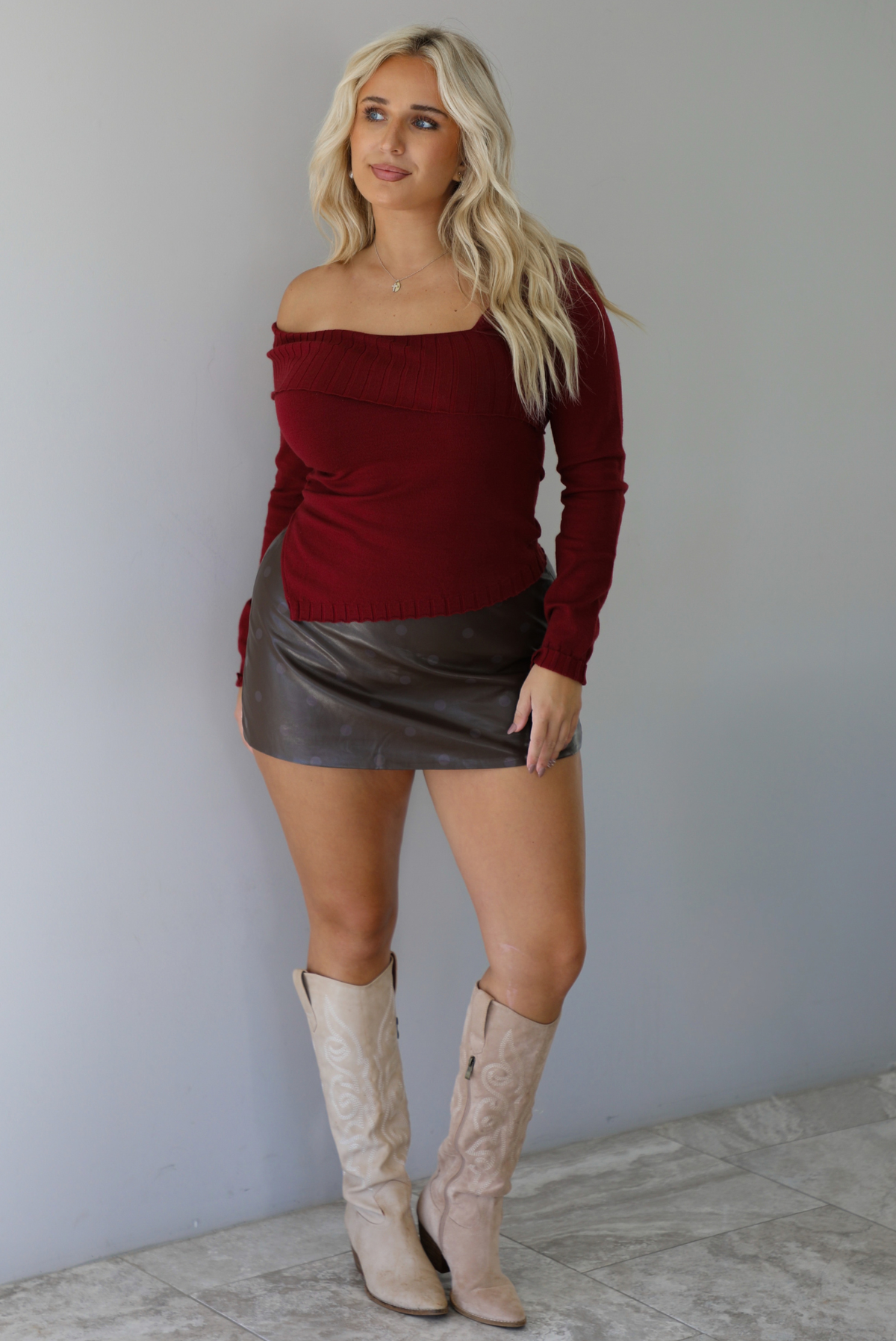 Woman wearing a maroon knit, long sleeved, off the shoulder knit top paired with a dark brown vegan leather mini skirt with a monochromatic polka dot print pattern, and beige knee high boots, standing indoors against a white wall with white marble flooring. 