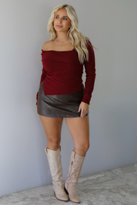 Woman wearing a maroon knit, long sleeved, off the shoulder knit top paired with a dark brown vegan leather mini skirt with a monochromatic polka dot print pattern, and beige knee high boots, standing indoors against a white wall with white marble flooring. 